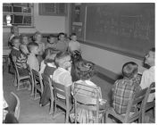 Children Sitting in a Circle of Chairs Facing the Blackboard